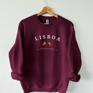 May include: Burgundy sweatshirt with white text that reads "LISBOA PORTUGAL 38.7223&deg; N, 9.1393&deg; W" and two crossed Portuguese flags.