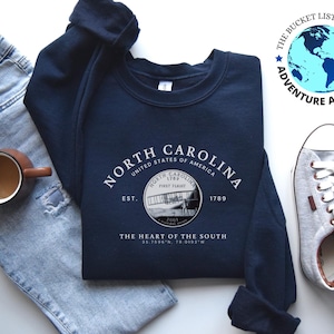 May include: A navy blue sweatshirt with a white graphic design featuring the text "NORTH CAROLINA" and a vintage airplane image. The text "UNITED STATES OF AMERICA" is also included, along with the text "EST. 1789" and "THE HEART OF THE SOUTH". The coordinates "35.7500&deg;N, 79.0193&deg;W" are also included in the design.