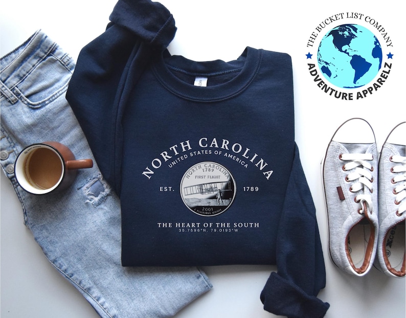 May include: A navy blue sweatshirt with a white graphic design featuring the text "NORTH CAROLINA" and a vintage airplane image. The text "UNITED STATES OF AMERICA" is also included, along with the text "EST. 1789" and "THE HEART OF THE SOUTH". The coordinates "35.7500&deg;N, 79.0193&deg;W" are also included in the design.