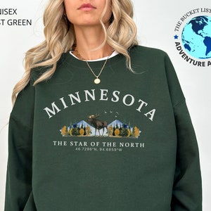 K&ouml;nnte beinhalten: Gr&uuml;n-farbener Sweatshirt mit einem wei&szlig;en Grafikdruck eines Elchs vor einer Bergkette mit B&auml;umen. Der Text "MINNESOTA" ist in einem wei&szlig;en Bogen &uuml;ber dem Elch gedruckt. Der Text "THE STAR OF THE NORTH" ist unter dem Elch gedruckt, zusammen mit den Koordinaten "46.7296'N, 94.6859'W".