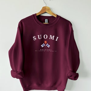 以下が含まれることがあります： ワインレッドのトレーナーに、白で「SUOMI」と「HELSINKI」の文字が書かれており、2つのフィンランド国旗が交差したグラフィックが描かれています。テキストの下には「60.1699&deg; N, 24.9384&deg; E」の座標が印刷されています。