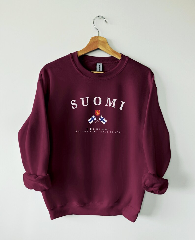 以下が含まれることがあります： ワインレッドのトレーナーに、白で「SUOMI」と「HELSINKI」の文字が書かれており、2つのフィンランド国旗が交差したグラフィックが描かれています。テキストの下には「60.1699&deg; N, 24.9384&deg; E」の座標が印刷されています。