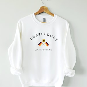 Op de afbeelding: Witte trui met zwarte tekst die "DÜSSELDORF" zegt met twee gekruiste Duitse vlaggen en de tekst "DEUTSCHLAND 51.2277° N, 6.7735° E" eronder.