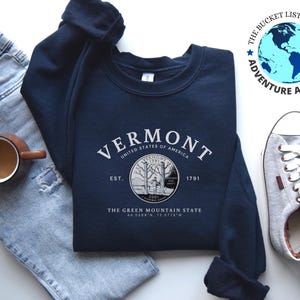 May include: A navy blue sweatshirt with a white graphic design featuring the text "VERMONT", "UNITED STATES OF AMERICA", "EST. 1791", "THE GREEN MOUNTAIN STATE", and coordinates. The graphic also includes a vintage-style image of a tree.