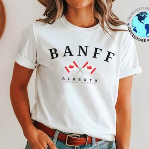 Pode incluir: Camiseta branca com texto preto que diz "BANFF" e "ALBERTA" com duas bandeiras canadenses cruzadas e coordenadas. A camiseta faz parte da marca "The Bucket List Company Adventure Apparel".