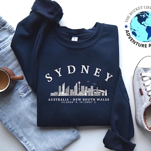 May include: A navy blue sweatshirt with a white graphic of the Sydney skyline and the text "SYDNEY, AUSTRALIA - NEW SOUTH WALES 33.8688° S, 151.2093° E".