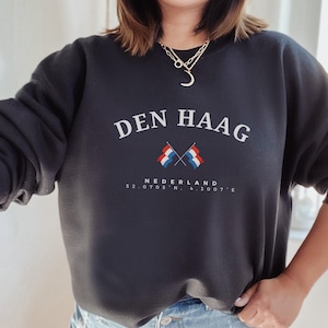 May include: Black sweatshirt with white text that reads "DEN HAAG" with two crossed Dutch flags underneath. The text "NEDERLAND" is below the flags, and the coordinates "52.0705° N, 4.3007° E" are below that.