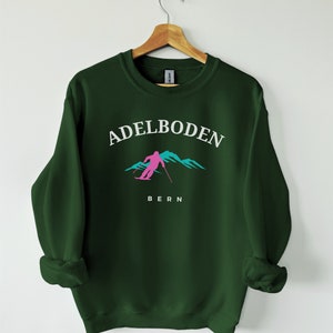 Op de afbeelding: Een donkergroen sweatshirt met een witte afbeelding van een skiër voor bergen. De tekst "Adelboden" staat boven de afbeelding en "Bern" staat onder de afbeelding.