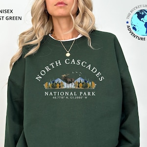 May include: A forest green unisex sweatshirt featuring the text "NORTH CASCADES NATIONAL PARK" and coordinates. The design includes a moose, trees, and mountains. The upper left corner reads "UNISEX FOREST GREEN". A logo for "THE BUCKET LIST COMPANY" is in the upper right.