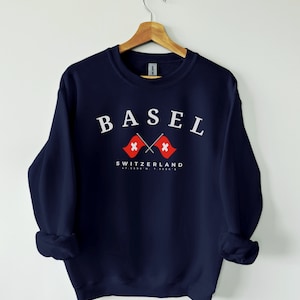 Peut inclure: Un sweat-shirt bleu marine avec le texte "BASEL" en blanc, deux drapeaux suisses croisés et le texte "SWITZERLAND 47.5586° N, 7.5886° E" en blanc.