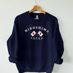 Puede incluir: Sudadera azul marino con un gráfico blanco de dos banderas japonesas cruzadas con el texto "Hiroshima Japón 34.3955° N, 132.4555° E" debajo.