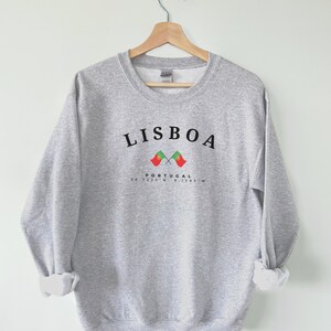 May include: Grey sweatshirt with the text "LISBOA" in black, two crossed Portuguese flags, and the text "PORTUGAL 38.7223&deg;N 9.1393&deg;W" in black.