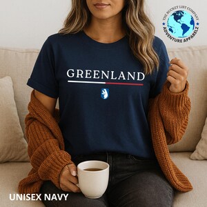 Pode incluir: Camiseta azul marinho com a palavra "GREENLAND" em branco, uma faixa branca e vermelha e um pequeno emblema. A camiseta tamb&eacute;m tem o texto "UNISEX NAVY" na parte inferior. Parte da cole&ccedil;&atilde;o "Adventure Apparelz".