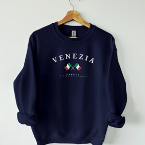 May include: Navy blue sweatshirt with the word "VENEZIA" in white arched text above two crossed Italian flags. Below the flags, the word "VENETO" and coordinates are printed in white. The sweatshirt is hanging on a wooden hanger.