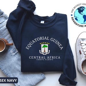Puede incluir: Sudadera azul marino con el texto "EQUATORIAL GUINEA CENTRAL AFRICA" y un escudo de armas. La sudadera también tiene el texto "UNISEX NAVY". La sudadera se muestra con vaqueros, una taza y zapatillas.