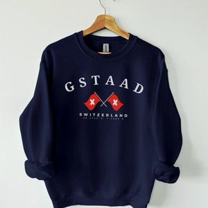 Op de afbeelding: Een donkerblauwe trui met een witte afbeelding van de tekst "GSTAAD" met twee gekruiste Zwitserse vlaggen en de tekst "SWITZERLAND" eronder. De coördinaten "46.4745°N, 7.2854°E" zijn onder de tekst gedrukt.