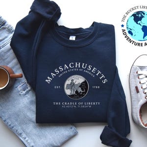 Puede incluir: Sudadera azul marino con la palabra "MASSACHUSETTS" en arco. Debajo, un gráfico circular con un sello estatal y el texto "THE CRADLE OF LIBERTY". La imagen también muestra vaqueros, una taza de café y zapatillas grises.