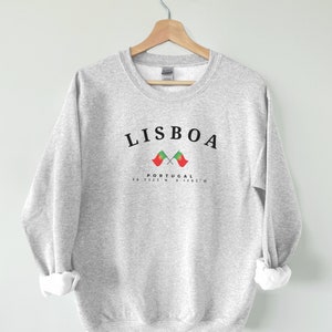 May include: Grey sweatshirt with the text "LISBOA" in black, two crossed Portuguese flags, and the text "PORTUGAL 38.7223&deg;N 9.1393&deg;W" in black.