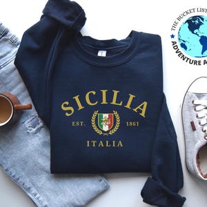 Könnte beinhalten: Marineblaues Sweatshirt mit goldfarbenem "SICILIA" Schriftzug, Wappen und "ITALIA" darunter. Das Bild zeigt eine hellblaue Jeans, graue Turnschuhe und eine Kaffeetasse. Das Logo von The Bucket List Company ist sichtbar.