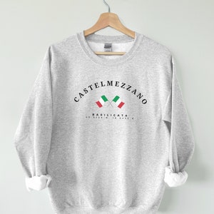 May include: A gray sweatshirt with the text "CASTELMEZZANO BASILICATA 40.5286 N 16.0455 E" and two crossed Italian flags printed on the front.
