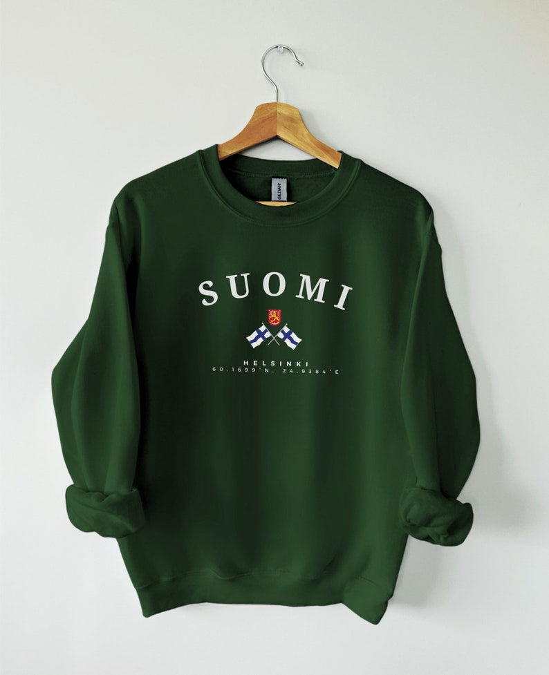以下が含まれることがあります： 白い文字で「SUOMI」と書かれた濃い緑色のスウェットシャツ。フィンランドの国旗が2つ交差しており、白い文字で「HELSINKI 60.1699&deg; N, 24.9384&deg; E」と書かれている。