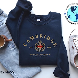 May include: A navy blue sweatshirt with "CAMBRIDGE" arched across the front in gold lettering. Below the text is a crest with the United Kingdom flag and the text "EST 1209". The sweatshirt is paired with jeans, a coffee mug, and gray sneakers.