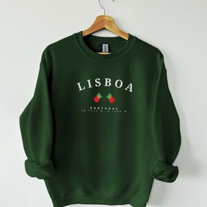 May include: A dark green sweatshirt with the word "LISBOA" printed in white on the front. Below the word is a small image of two crossed Portuguese flags and the text "PORTUGAL 38.7225&deg;N, 9.1393&deg;W".