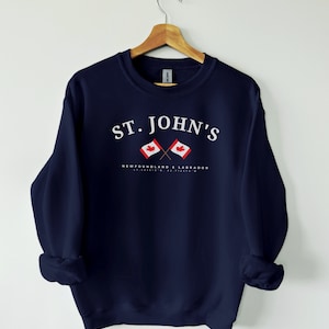 May include: Navy blue sweatshirt with the text "ST. JOHN'S" in white, above two crossed Canadian flags. Below the flags, the text "NEWFOUNDLAND & LABRADOR" and coordinates are printed in white. The sweatshirt is hanging on a wooden hanger.