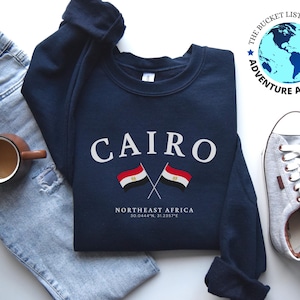 May include: A navy blue sweatshirt with the word "CAIRO" printed in white, along with two crossed flags of Egypt. The text "NORTHEAST AFRICA" and the coordinates "30.0444°N, 31.2357°E" are printed below the flags.