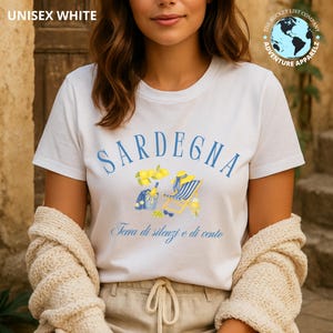 May include: White unisex t-shirt with the word "SARDEGNA" in blue arched text. Below the text is a watercolor illustration of lemons, a beach chair, and a bucket. The text "Terra di silenzi e di vento" is below the illustration.