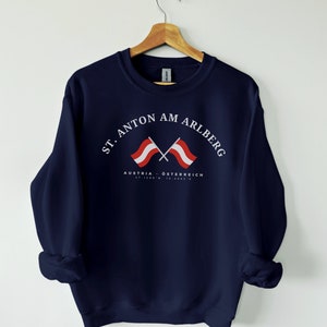 May include: Navy blue sweatshirt with a white graphic of two crossed Austrian flags and the text "St. Anton am Arlberg, Austria - Österreich, 47.1208° N, 10.2483° E".