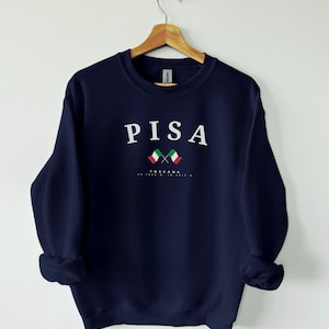 Può includere: Una felpa blu navy con la scritta "PISA" in lettere bianche. Sotto il testo ci sono due bandiere italiane incrociate e il testo "TOSCANA 43.7202° N, 10.4017° E".