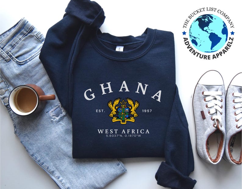 Puede incluir: Sudadera azul marino con la palabra "GHANA" en letras blancas arqueadas. Debajo, un escudo, el texto "EST. 1957" y "WEST AFRICA" con coordenadas. La sudadera est&aacute; junto a zapatillas grises y unos vaqueros azules.