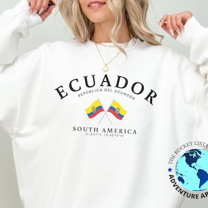 May include: White sweatshirt with black text that reads "ECUADOR" and "REPÚBLICA DEL ECUADOR". Below the text are two crossed flags of Ecuador. The text "SOUTH AMERICA" and coordinates "0.1807°S, 78.4678°W" are below the flags. A blue and white globe with the text "THE BUCKET LIST COMPANY" and "ADVENTURE APPARELZ" is in the lower right corner.