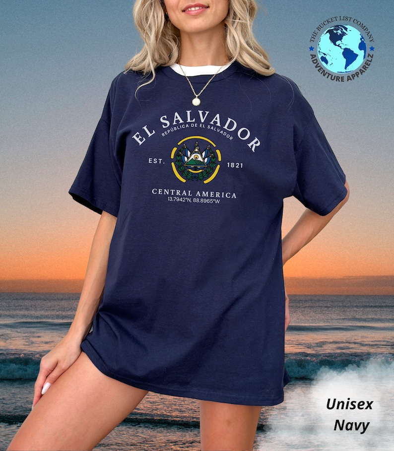 May include: A navy blue unisex t-shirt featuring a white graphic of the El Salvador coat of arms, the text "El Salvador", "Republica de El Salvador", "Est. 1821", "Central America", and the coordinates "13.7942&deg;N 88.8965&deg;W".