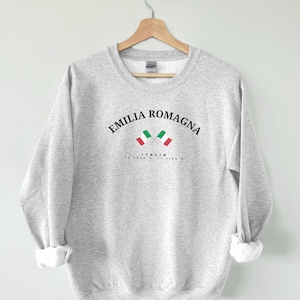 Op de afbeelding: Grijs sweatshirt met de tekst "Emilia Romagna" en twee gekruiste Italiaanse vlaggen met de tekst "ITALIA" en coördinaten eronder.
