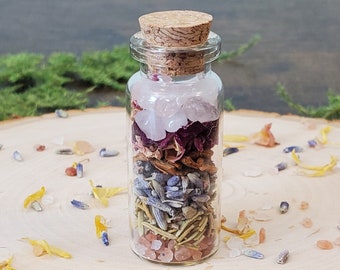 May include: A small glass bottle filled with layers of dried flowers, herbs, and crystals. The bottle has a cork stopper and is sitting on a wooden surface.