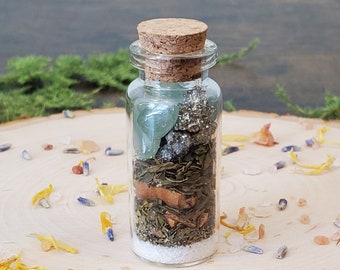 May include: A small glass bottle with a cork stopper filled with green and brown herbs, crystals, and white salt. The bottle is sitting on a wooden surface with scattered dried flowers.