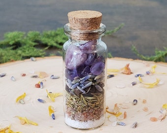 May include: A small glass bottle with a cork stopper filled with amethyst crystals, lavender, herbs, and salt. The bottle is sitting on a wooden surface with dried flower petals scattered around it.
