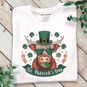 Peut inclure: T-shirt blanc avec un motif de vache des Highlands portant un chapeau haut-de-forme vert, des lunettes en forme de cœur et un trèfle dans la bouche. Le design comprend des trèfles et le texte "St. Patrick's Day" sur une bannière verte.