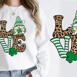 Op de afbeelding: Witte sweatshirt met een kleurrijke "LOVE" print. De letters "L" en "E" hebben een luipaardprint, terwijl de "V" groen en wit gestreept is. Een kabouter met een groene hoed en een klavertjevier is afgebeeld.