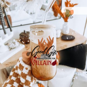 Könnte beinhalten: Ein Glasbecher mit einem Holzdeckel und einem Strohhalm. Der Becher enthält eine braune Flüssigkeit und eine Grafik mit dem Text "Good Girl Always Falling for the Villain".