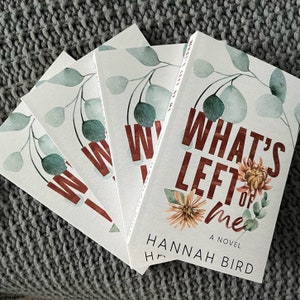 May include: Four copies of the novel "What's Left of Me" by Hannah Bird are arranged on a gray knitted surface. The book covers feature a floral design with the title in a rust-colored font and a watercolor illustration of leaves and flowers.