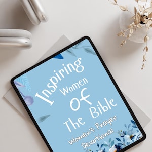 Peut inclure: Une tablette numérique affiche le texte "Inspiring Women of the Bible" sur un fond bleu clair avec des motifs floraux. La tablette est posée sur une surface blanche, avec un casque argenté et une composition florale décorative en arrière-plan.