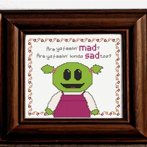 May include: Cross-stitch pattern featuring a green cartoon character with a pink dress and the text "Are ya feelin' mad? Are ya feelin' kinda sad too?"