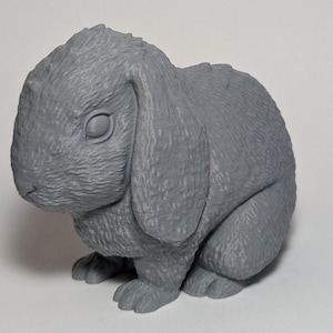 May include: A gray, three-dimensional rabbit figurine with floppy ears. The bunny is in a seated position, with detailed fur texture and closed eyes. The figurine is made of a solid, matte material.