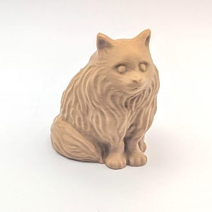 May include: A tan cat figurine in a seated pose. The cat has long, textured fur and a detailed face with small, round eyes and pointed ears. The figurine is made of a solid material and sits on a white surface.