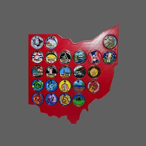 May include: A red Ohio-shaped display board holds a collection of colorful round coins. Each coin features unique designs and text, including images of landmarks and illustrations. The coins are arranged in rows and columns on the board.
