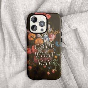 May include: A black phone case with a floral design and the text "COME WHAT MAY". The design features a dark background with orange, red, pink, and blue flowers. The phone case is lying on a white fabric.