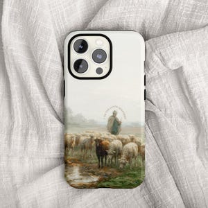May include: A white phone case with a pastoral scene featuring a shepherd and a flock of sheep. The case has a black border and a camera cutout. The text "I AM THE GOOD SHEPHERD" is printed on the case.